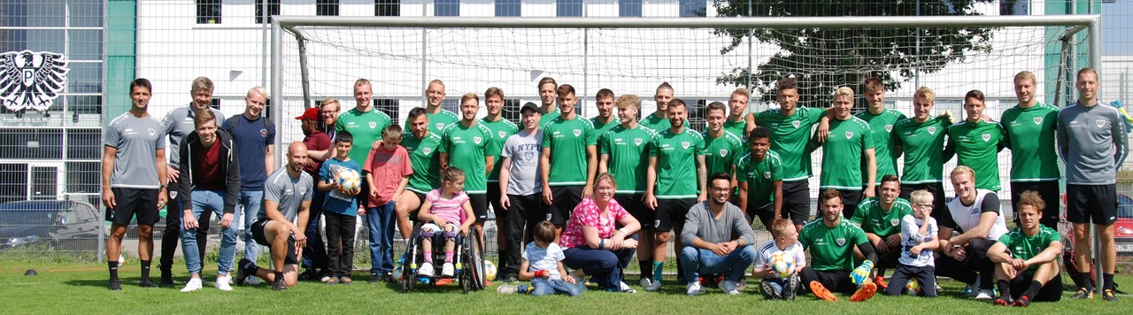 Die Mannschaft und Trainer des SC Preußen Münster posieren in Trainingskleidung mit den Teilnehmer_innen der Ferienfreizeit sowie den Betreuer_innen für ein Gruppenphoto vor einem Fußballtor.